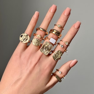 Può includere: Una collezione di anelli color oro indossati sulle dita, caratterizzati da vari design tra cui medaglioni circolari stile moneta, una placca rettangolare con motivo a serpente, una pietra quadrata bianca, fasce lisce e anelli con piccoli cristalli.