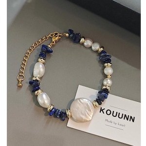 May include: A bracelet featuring irregular white freshwater pearls, small deep blue lapis lazuli stone chips, and gold-toned metal spacers. The piece includes a gold-toned chain extender and a white card with the text KOUUNN Made by heart.