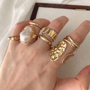 Può includere: Una collezione di anelli color oro indossati sulle dita. Un anello presenta una grande perla bianca irregolare. Altri anelli includono una fascia liscia, una fascia ondulata, un anello largo con superficie martellata e una fascia aperta.