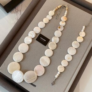May include: A necklace and bracelet set featuring circular white mother-of-pearl beads of varying sizes. The jewelry includes silver-toned metal clasps and extender chains. A small black rectangular tag with the white text KOUUNN is included.