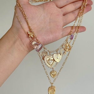 May include: A multi-layered gold-tone chain necklace featuring various charms including a sun, a horseshoe, and several heart-shaped pendants. Some hearts are metallic gold, while others are translucent purple glass with a faceted finish.