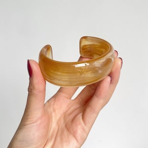 May include: A wide, open-ended cuff bracelet made of polished resin in a translucent amber color. The material features a swirling, marbled pattern of light and dark golden-brown tones throughout the smooth, rounded surface of the circular band.