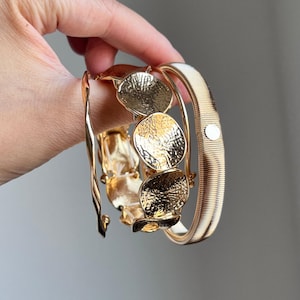 May include: A set of three gold-toned bracelets. One bracelet features a twisted wire design, the second consists of linked circular discs with a hammered texture, and the third is a wide, ribbed band with a small circular accent. Text reads CEISUR.