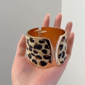 May include: A gold-toned metal cuff bracelet featuring a textured leopard print pattern with faux fur detailing. The bracelet has a hinged opening mechanism and a smooth, polished gold interior finish, showcasing a wide, open-ended design.