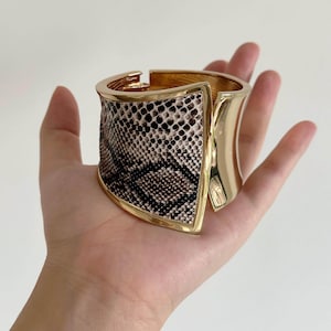 May include: A wide hinged cuff bracelet featuring a polished gold-tone metal frame and a central panel with a brown and black snakeskin pattern. The bracelet has a curved, architectural shape with a smooth metallic finish on the sides and back.