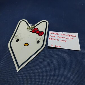 May include: A white Hello Kitty-shaped sticker with a red bow, black outline, and a yellow oval nose. The sticker has two black oval eyes and a metal fastener. A handwritten note reads "VARIASI TEM PELAN KACA HELLO KITTY BENTUK LOVE" and "K 295".