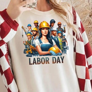 Può includere: T-shirt color crema con grafica del Labor Day con un gruppo di operai edili e la scritta "LABOR DAY". Il design include una donna con un casco giallo e una tuta blu, che tiene in mano un casco giallo. Altri operai indossano caschi e abiti da lavoro.