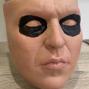 Könnte beinhalten: Realistische Maske eines menschlichen Gesichts mit schwarzem Augen-Make-up. Die Maske ist hautfarben mit einer grauen Kopfbedeckung. Das Gesicht hat einen neutralen Ausdruck, mit sichtbaren Falten und detaillierten Merkmalen.