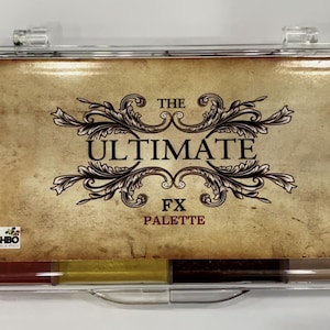 Puede incluir: Un estuche de plástico transparente que contiene una paleta de maquillaje. La paleta presenta un diseño de estilo vintage con las palabras "THE ULTIMATE FX PALETTE" en negro. La paleta incluye varios bloques de maquillaje de colores.