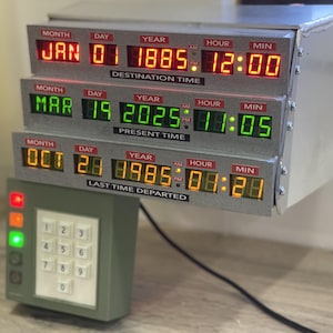 Może przedstawiać: Urządzenie wyświetlające obwód czasowy z trzema poziomymi rzędami cyfrowych odczytów LED oznaczonych jako Destination Time, Present Time i Last Time Departed, w kolorze czerwonym, zielonym i żółtym, oraz oddzielna klawiatura z numerowanymi przyciskami.