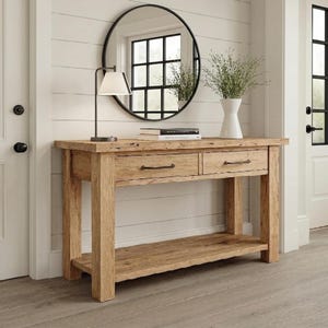 May include: A rustic wooden console table with two drawers and a lower shelf. A black-framed round mirror hangs above the table, reflecting a window. A white lamp and a white vase with greenery sit on the table. The table is made of light-colored wood.