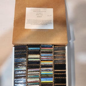 May include: A cardboard box containing numerous small, rectangular glass samples in various colors. The samples are arranged in vertical rows within the box. A white label with text is visible inside the box.
