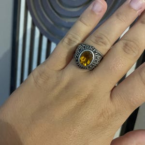 Op de afbeelding: Een zilverkleurige metalen ring met een grote, ovaal geslepen barnsteenkleurige edelsteen in het midden. De ringband is gegraveerd met de tekst CLARKFIELD en bevat kleine steraccenten rondom de zetting van de centrale steen.