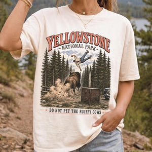 May include: An ivory short-sleeve t-shirt featuring a graphic print of Yellowstone National Park. The design shows a person being tossed by a bison in a forest setting, with the text "YELLOWSTONE NATIONAL PARK" and "DO NOT PET THE FLUFFY COWS".