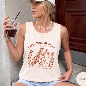 May include: An ivory sleeveless tank top featuring a rust-coloured graphic print of a praying mantis surrounded by floral motifs and the text "GIRLS WILL BE GIRLS" arched above the insect. The design includes intricate patterns on the mantis body.