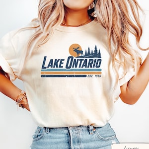 May include: An ivory t-shirt featuring a graphic of a moose silhouette, pine trees, and a sun above the text LAKE ONTARIO and EST. 1615. The design includes horizontal stripes in blue and orange tones, printed on the center of the chest.