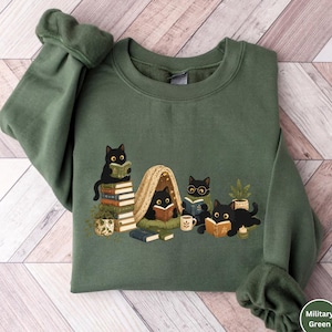 Puede incluir: Una sudadera de cuello redondo color verde militar con una ilustraci&oacute;n gr&aacute;fica de cuatro gatos negros leyendo libros. Los gatos est&aacute;n situados alrededor de una pila de libros, una peque&ntilde;a tienda de campa&ntilde;a, una planta, una taza y una vela.