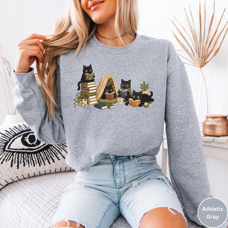 Puede incluir: Una sudadera gris jaspeado de cuello redondo con un estampado gr&aacute;fico de cuatro gatos negros leyendo libros. La ilustraci&oacute;n muestra gatos sentados sobre pilas de libros, dentro de una peque&ntilde;a tienda de campa&ntilde;a y tumbados con libros abiertos.