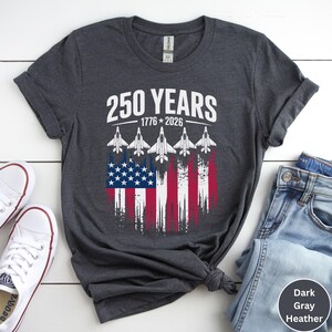May include: Dark gray heather t-shirt with the text "250 YEARS 1776-2026" above a graphic of the American flag with jets. The shirt is paired with blue jeans and white sneakers.