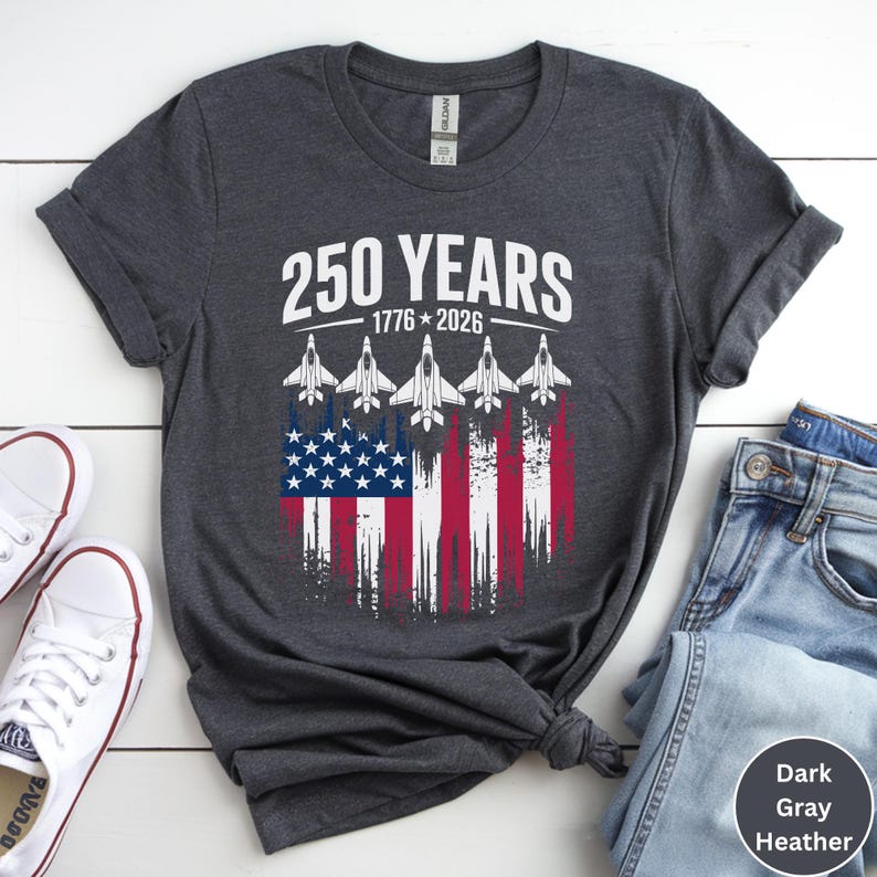 May include: Dark gray heather t-shirt with the text "250 YEARS 1776-2026" above a graphic of the American flag with jets. The shirt is paired with blue jeans and white sneakers.