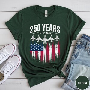 May include: Forest green t-shirt with the text "250 YEARS 1776-2026" above a graphic of the American flag and five fighter jets. White sneakers and blue jeans are visible.