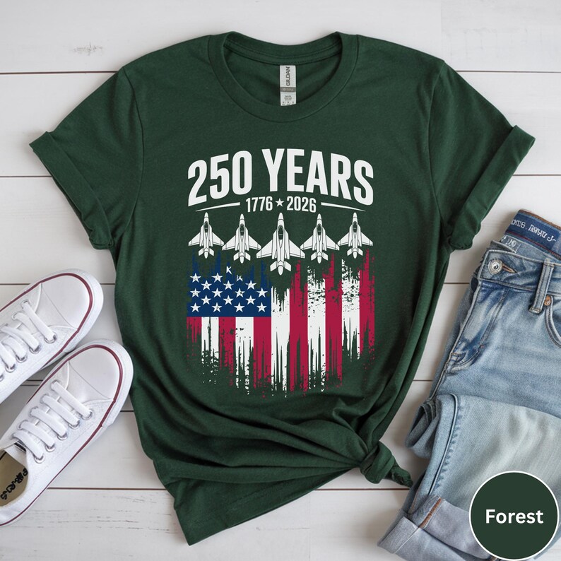 May include: Forest green t-shirt with the text "250 YEARS 1776-2026" above a graphic of the American flag and five fighter jets. White sneakers and blue jeans are visible.