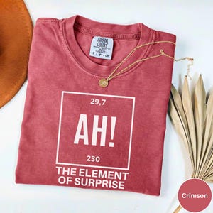 May include: Crimson t-shirt featuring the text "AH! THE ELEMENT OF SURPRISE" in a white box. A gold necklace with a pendant is on the shirt. The shirt has a Comfort Colors label.