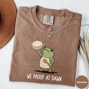 May include: Espresso-colored t-shirt with a graphic of a green frog wearing an apron and holding a bread bowl. The text "WE PROOF AT DAWN" is printed below the frog. A silver necklace with a pendant is draped on the shirt.