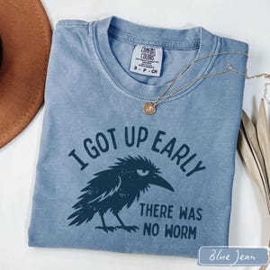 May include: A blue short-sleeved t-shirt featuring a dark blue graphic of a grumpy crow with the text I GOT UP EARLY THERE WAS NO WORM. The shirt has a Comfort Colors brand label at the inner neckline and is paired with a gold-toned chain necklace.