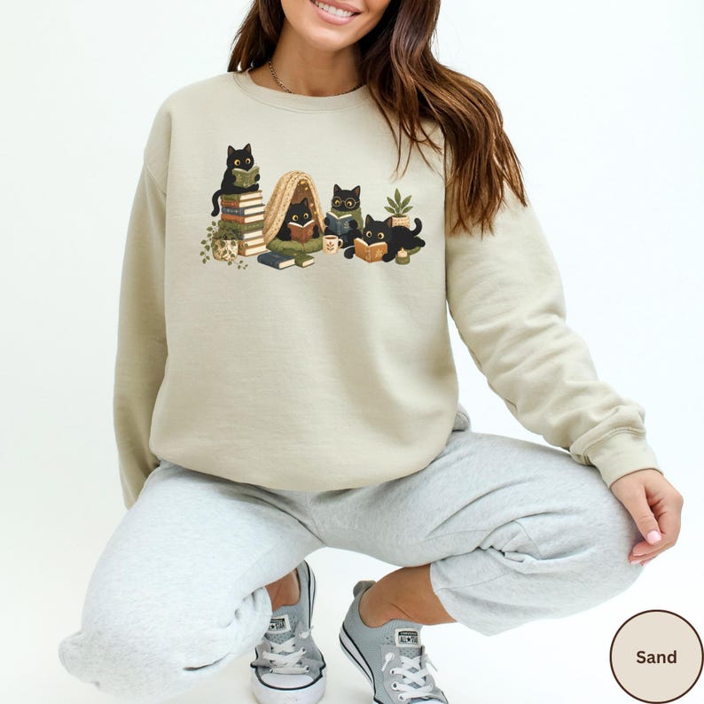 Puede incluir: Una sudadera de cuello redondo color arena con un estampado gr&aacute;fico de cuatro gatos negros leyendo libros. La ilustraci&oacute;n incluye gatos sentados sobre pilas de libros, dentro de una peque&ntilde;a tienda de campa&ntilde;a y cerca de una planta en maceta.