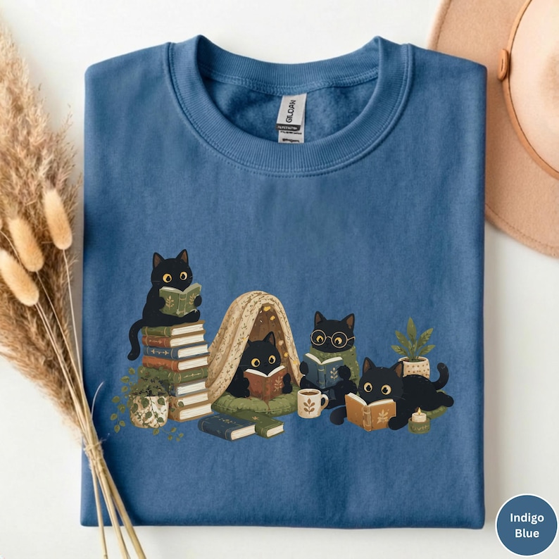 Puede incluir: Una sudadera de cuello redondo color azul &iacute;ndigo con una ilustraci&oacute;n gr&aacute;fica de cuatro gatos negros leyendo libros. Los gatos est&aacute;n situados alrededor de una peque&ntilde;a tienda de campa&ntilde;a, pilas de libros, una planta, una taza y una vela.