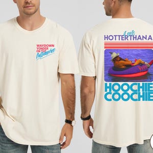 May include: Ivory-colored t-shirt with text on the front and back. The front reads "WAYDOWN YONDER ON THE Chattanooga". The back features a graphic of a person in a tube with the text "it gets HOTTER THANA HOOCHIE COOCHIE".