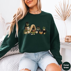 Puede incluir: Una sudadera de manga larga color verde bosque con una ilustraci&oacute;n gr&aacute;fica de cuatro gatos negros leyendo libros. Los gatos est&aacute;n situados alrededor de una peque&ntilde;a tienda de mantas, pilas de libros, una planta, una taza y una vela.