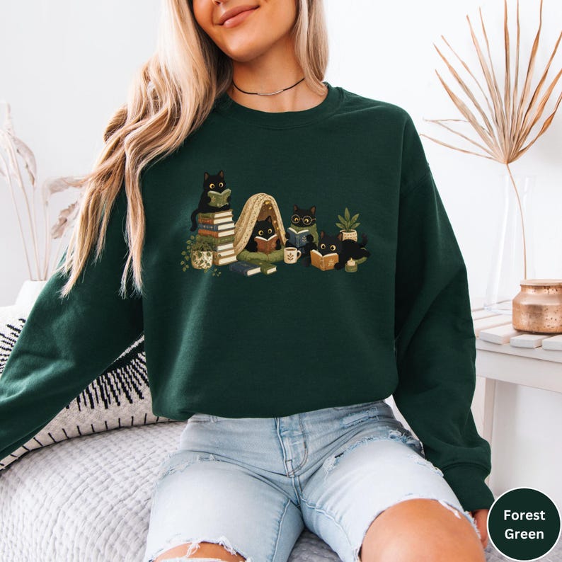 Puede incluir: Una sudadera de manga larga color verde bosque con una ilustraci&oacute;n gr&aacute;fica de cuatro gatos negros leyendo libros. Los gatos est&aacute;n situados alrededor de una peque&ntilde;a tienda de mantas, pilas de libros, una planta, una taza y una vela.