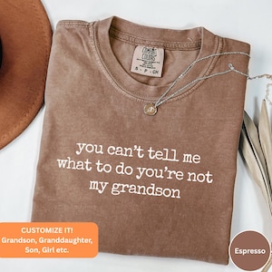 May include: Espresso-colored t-shirt with the text "you can't tell me what to do you're not my grandson" in white. The shirt has a crew neck and short sleeves. A silver necklace with a pendant is draped across the shirt.