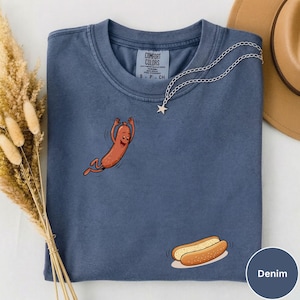 May include: Denim blue t-shirt with a cartoon hot dog design. The shirt features a smiling hot dog character and a hot dog bun. A silver necklace with a star pendant is draped on the shirt. The tag reads "COMFORT COLORS".