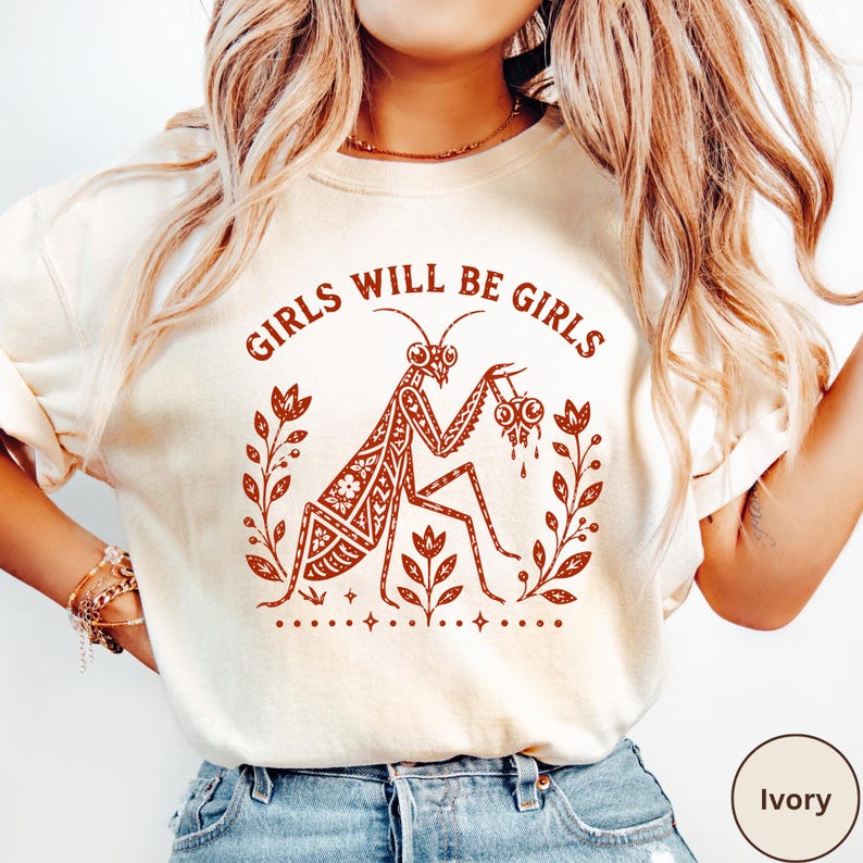 May include: An ivory t-shirt featuring a rust-coloured graphic of a praying mantis surrounded by floral elements and the text GIRLS WILL BE GIRLS. The design includes intricate patterns on the insect and small star accents at the bottom.