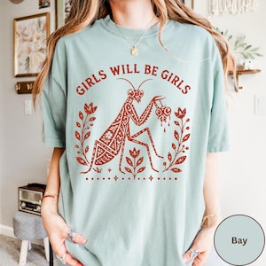 May include: A light sage green t-shirt featuring a rust-coloured graphic print of a praying mantis holding a heart. The text GIRLS WILL BE GIRLS is arched above the insect, which is flanked by floral stems and small star accents on the front.