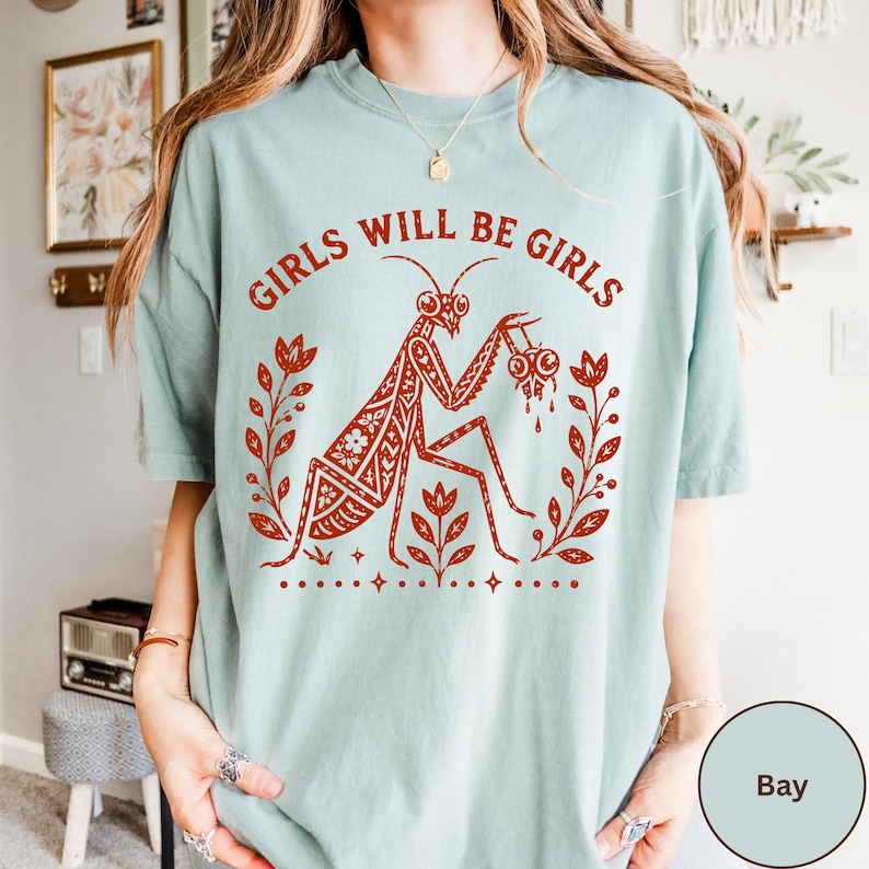 May include: A light sage green t-shirt featuring a rust-coloured graphic print of a praying mantis holding a heart. The text GIRLS WILL BE GIRLS is arched above the insect, which is flanked by floral stems and small star accents on the front.