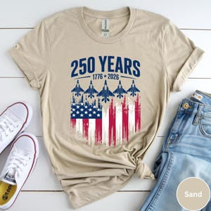 May include: A sand-colored t-shirt featuring the text "250 YEARS 1776-2026" in blue. Below the text is an American flag design with jets flying upwards. White sneakers and blue jeans are visible.
