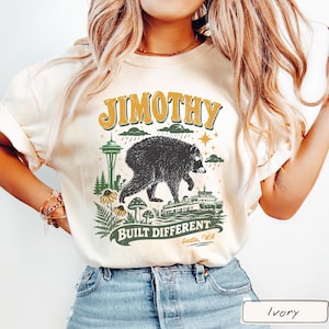 May include: An ivory t-shirt featuring a graphic print with the text JIMOTHY and BUILT DIFFERENT Seattle, WA. The design includes a black bear, the Space Needle, a ferry boat, mushrooms, ferns, and rain clouds in shades of green, yellow, and black.