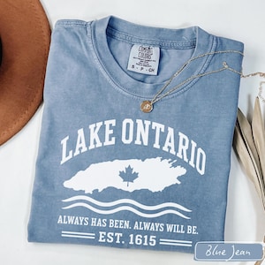 May include: A blue short-sleeve t-shirt with a white graphic featuring the text LAKE ONTARIO, a maple leaf icon, and the phrase ALWAYS HAS BEEN. ALWAYS WILL BE. EST. 1615. The shirt has a Comfort Colors brand label at the inner neckline.
