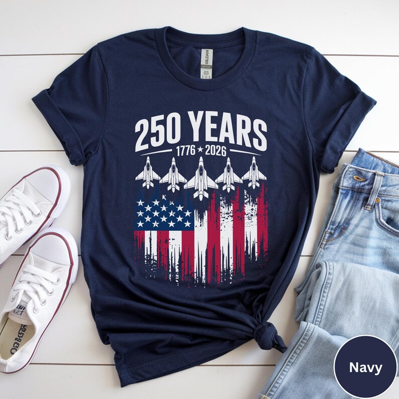 May include: Navy blue t-shirt featuring the text "250 YEARS 1776-2026" above a graphic of the American flag with jets. The design is in white, red, and blue. White sneakers and blue jeans are also visible.