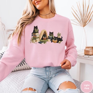Puede incluir: Una sudadera de cuello redondo en color rosa claro con una ilustraci&oacute;n gr&aacute;fica de cinco gatos negros leyendo libros. Los gatos aparecen sentados sobre pilas de libros, dentro de una peque&ntilde;a tienda de campa&ntilde;a y tumbados leyendo, junto a una planta.