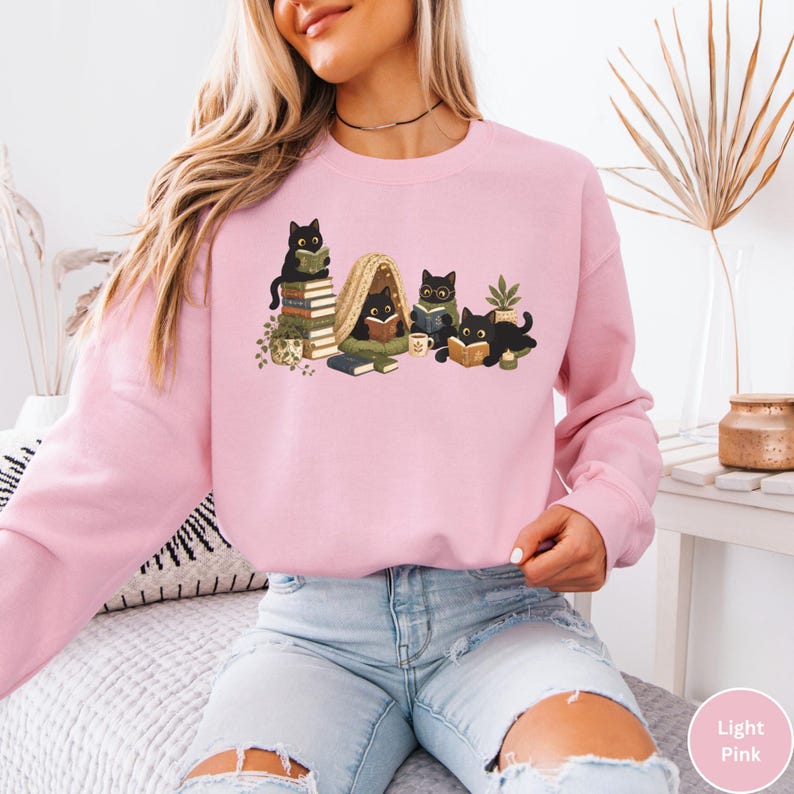 Puede incluir: Una sudadera de cuello redondo en color rosa claro con una ilustraci&oacute;n gr&aacute;fica de cinco gatos negros leyendo libros. Los gatos aparecen sentados sobre pilas de libros, dentro de una peque&ntilde;a tienda de campa&ntilde;a y tumbados leyendo, junto a una planta.