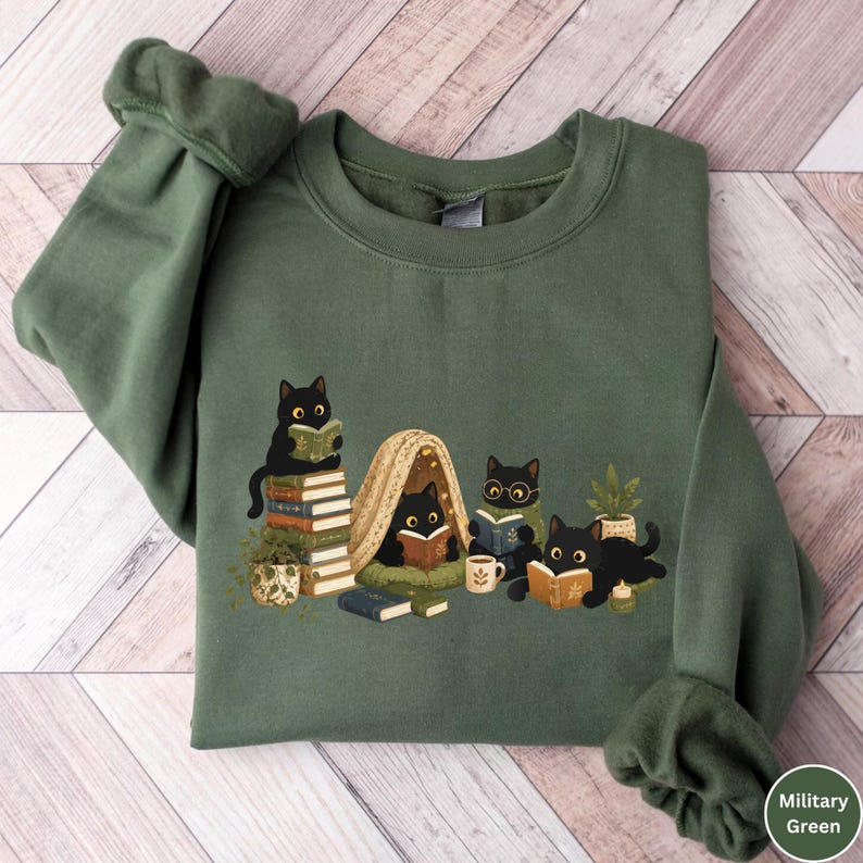 Puede incluir: Una sudadera de cuello redondo color verde militar con una ilustraci&oacute;n gr&aacute;fica de cuatro gatos negros leyendo libros. Los gatos est&aacute;n situados alrededor de una pila de libros, una peque&ntilde;a tienda de campa&ntilde;a, una planta, una taza y una vela.