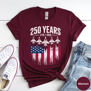 May include: Maroon t-shirt with the text "250 YEARS 1776-2026" and an American flag design. The design includes five white fighter jets. The shirt is paired with white sneakers and blue jeans.