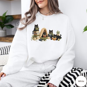 Puede incluir: Una sudadera blanca de manga larga con una ilustraci&oacute;n gr&aacute;fica de cuatro gatos negros leyendo libros. Los gatos est&aacute;n situados alrededor de una peque&ntilde;a tienda de campa&ntilde;a, pilas de libros, una planta en maceta y una taza, en tonos verdes y marrones.