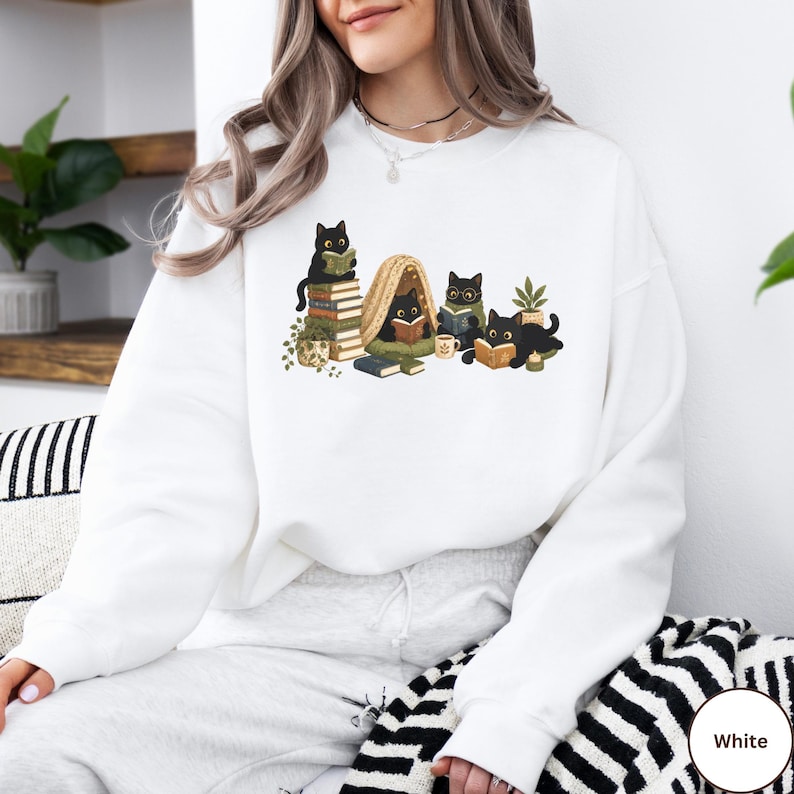 Puede incluir: Una sudadera blanca de manga larga con una ilustraci&oacute;n gr&aacute;fica de cuatro gatos negros leyendo libros. Los gatos est&aacute;n situados alrededor de una peque&ntilde;a tienda de campa&ntilde;a, pilas de libros, una planta en maceta y una taza, en tonos verdes y marrones.