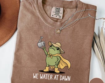 Peut inclure: T-shirt couleur expresso avec une grenouille de dessin animé portant un chapeau et une cape, tenant un arrosoir. Le texte "WE WATER AT DAWN" est imprimé en dessous. Un collier argenté avec un pendentif est posé sur le t-shirt.